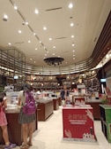 A bookstore interior with high ceilings adorned with numerous spotlights. Shelves filled with a variety of books line the walls, reaching up to the ceiling. Several wooden tables in the center display an assortment of books and advertisements. People are browsing the books, with a focus on a person wearing a backpack close to the foreground. The atmosphere is calm and inviting, with a well-organized layout.