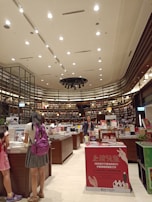 A bookstore interior with high ceilings adorned with numerous spotlights. Shelves filled with a variety of books line the walls, reaching up to the ceiling. Several wooden tables in the center display an assortment of books and advertisements. People are browsing the books, with a focus on a person wearing a backpack close to the foreground. The atmosphere is calm and inviting, with a well-organized layout.