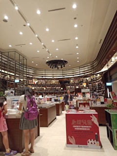 A bookstore interior with high ceilings adorned with numerous spotlights. Shelves filled with a variety of books line the walls, reaching up to the ceiling. Several wooden tables in the center display an assortment of books and advertisements. People are browsing the books, with a focus on a person wearing a backpack close to the foreground. The atmosphere is calm and inviting, with a well-organized layout.