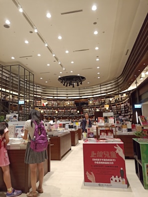 A bookstore interior with high ceilings adorned with numerous spotlights. Shelves filled with a variety of books line the walls, reaching up to the ceiling. Several wooden tables in the center display an assortment of books and advertisements. People are browsing the books, with a focus on a person wearing a backpack close to the foreground. The atmosphere is calm and inviting, with a well-organized layout.