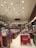 A bookstore interior with high ceilings adorned with numerous spotlights. Shelves filled with a variety of books line the walls, reaching up to the ceiling. Several wooden tables in the center display an assortment of books and advertisements. People are browsing the books, with a focus on a person wearing a backpack close to the foreground. The atmosphere is calm and inviting, with a well-organized layout.