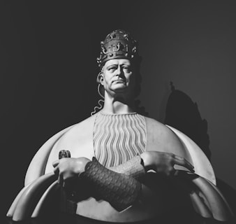 A monochrome photograph of a regal statue featuring a solemn male figure wearing an ornate crown. The figure is dressed in a detailed robe with a texture resembling waves. His arms are crossed with a firm and commanding presence, and his visage exudes authority and power. Shadows enhance the depth and drama of the sculpture.