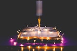An open book is placed on a surface with glowing string lights draped across it, casting a warm and inviting glow. Behind the book stands an illuminated candle on a holder, adding to the serene and contemplative atmosphere.