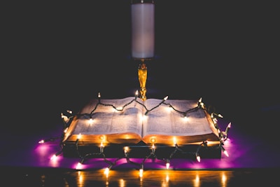 An open book is placed on a surface with glowing string lights draped across it, casting a warm and inviting glow. Behind the book stands an illuminated candle on a holder, adding to the serene and contemplative atmosphere.