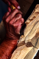 A pair of hands skillfully carves detailed patterns into a block of wood using a carving tool. The wood displays a series of intricate cuts and angled shapes, suggesting an ongoing artistic process. The focus on the hands and tool emphasizes craftsmanship and precision.