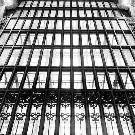 A large, intricate metal and glass window with multiple panes and ornate decorative elements at the bottom. The structure is symmetric, with vertical and horizontal bars creating rectangular sections. The light filters through, highlighting the design.