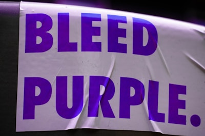 A white sticker with large, bold purple text that reads 'BLEED PURPLE.' The sticker appears to be slightly wrinkled and is placed against a dark background.