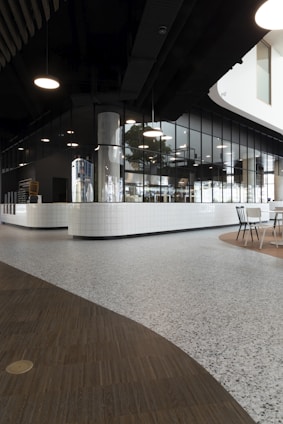 A modern interior space with sleek, dark ceilings and pendant lighting. The walls are composed of reflective glass windows and white tiled surfaces, creating a clean, minimalist aesthetic. The flooring is a mix of wood and terrazzo, adding texture to the environment. A seating area with chairs is visible in the corner, suggesting a social or dining space.