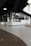 A modern interior space with sleek, dark ceilings and pendant lighting. The walls are composed of reflective glass windows and white tiled surfaces, creating a clean, minimalist aesthetic. The flooring is a mix of wood and terrazzo, adding texture to the environment. A seating area with chairs is visible in the corner, suggesting a social or dining space.