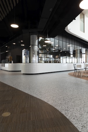 A modern interior space with sleek, dark ceilings and pendant lighting. The walls are composed of reflective glass windows and white tiled surfaces, creating a clean, minimalist aesthetic. The flooring is a mix of wood and terrazzo, adding texture to the environment. A seating area with chairs is visible in the corner, suggesting a social or dining space.