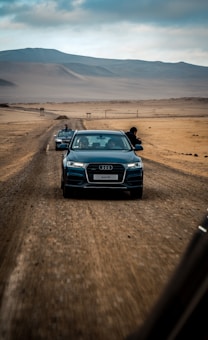 An Audi Q3 is driving along a rugged, desert road, followed by another vehicle in the background. The landscape features sandy terrain and distant hills under a cloudy sky. The scene captures an adventurous journey with a sense of motion.