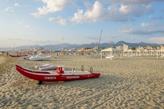 A red rescue boat labeled 'TERESITA SALVATAGGIO' is on a sandy beach. In the background, there are several other boats, beach chairs, umbrellas, and people. The sky is partly cloudy with soft light from the setting sun, and there are mountains visible in the distance.
