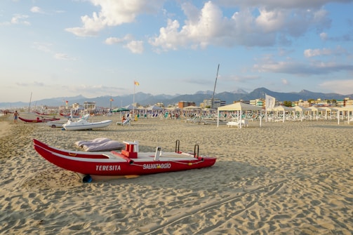 A red rescue boat labeled 'TERESITA SALVATAGGIO' is on a sandy beach. In the background, there are several other boats, beach chairs, umbrellas, and people. The sky is partly cloudy with soft light from the setting sun, and there are mountains visible in the distance.