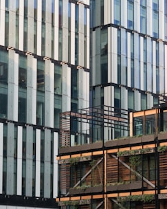 A modern architectural facade with a combination of glass and horizontal wooden elements. The building features large glass panels with vertical accents, and the lower section is adorned with wooden slats and greenery.
