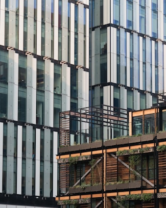 A modern architectural facade with a combination of glass and horizontal wooden elements. The building features large glass panels with vertical accents, and the lower section is adorned with wooden slats and greenery.
