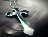 A metallic cross pendant on a beaded chain is lying on an open palm. The focus is on the cross, which has a shiny, reflective surface with curved edges. Soft lighting creates a gentle glow around the pendant, casting shadows and reflections.