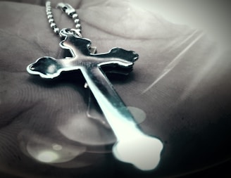 A metallic cross pendant on a beaded chain is lying on an open palm. The focus is on the cross, which has a shiny, reflective surface with curved edges. Soft lighting creates a gentle glow around the pendant, casting shadows and reflections.