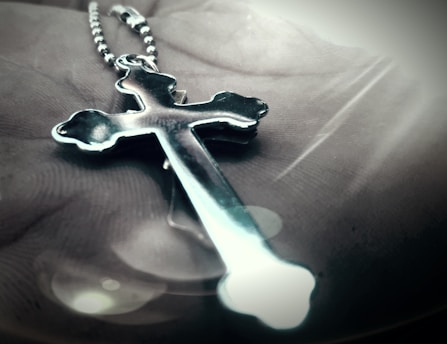 A metallic cross pendant on a beaded chain is lying on an open palm. The focus is on the cross, which has a shiny, reflective surface with curved edges. Soft lighting creates a gentle glow around the pendant, casting shadows and reflections.