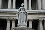 A stone statue of a regal figure dressed in ornate, historical clothing is seen holding a scepter and orb. The figure wears a crown and stands on a pedestal in front of a grand building with tall, classical columns.