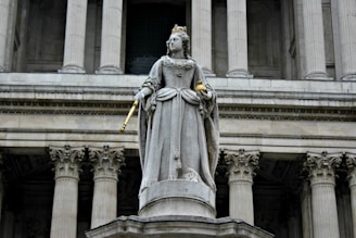 A stone statue of a regal figure dressed in ornate, historical clothing is seen holding a scepter and orb. The figure wears a crown and stands on a pedestal in front of a grand building with tall, classical columns.