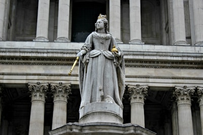 A stone statue of a regal figure dressed in ornate, historical clothing is seen holding a scepter and orb. The figure wears a crown and stands on a pedestal in front of a grand building with tall, classical columns.