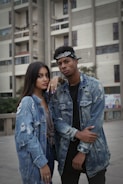 Two individuals stand confidently outside an urban building, both wearing distressed denim jackets. The background features a tall, modern building with balconies and windows. The people have a casual and stylish appearance.