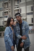 Two individuals stand confidently outside an urban building, both wearing distressed denim jackets. The background features a tall, modern building with balconies and windows. The people have a casual and stylish appearance.