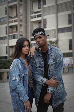 Two individuals stand confidently outside an urban building, both wearing distressed denim jackets. The background features a tall, modern building with balconies and windows. The people have a casual and stylish appearance.