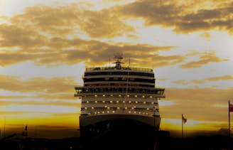 A large cruise ship is illuminated against a dramatic sky filled with dense clouds during sunset. The ship’s lights contrast with the dusk, highlighting its massive structure.