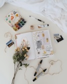 An artistic flat lay featuring an open sketchbook with various creative drawings and inspirational quotes. Surrounding the sketchbook are a palette of watercolor paints, a few brushes, dried flowers, a ball of twine, a few scattered pens, markers, a rubber stamp, and small print photos. The scene exudes a cozy and creative atmosphere, enhanced by the soft texture of a white fabric that partially covers part of the setup.
