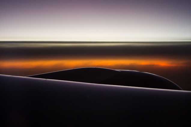 A dramatic sky with a gradient of colors ranging from dark grey to a fiery orange hue. The silhouette of a plane's engine or wing is visible against the backdrop, enhancing the sense of altitude and open space. The horizon is accentuated by the light transition, suggesting a sunrise or sunset.