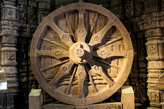 An intricately carved stone wheel with detailed sculptures and patterns on its surface, set against a backdrop of ornate stone architecture. The artwork includes relief sculptures of figures and decorative motifs, showing a high level of craftsmanship and cultural heritage.