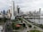 A modern cityscape featuring numerous tall skyscrapers with varied architectural designs. There are lush green parks, a winding road network, and a body of water reflecting some of the buildings.
