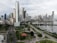A modern cityscape featuring numerous tall skyscrapers with varied architectural designs. There are lush green parks, a winding road network, and a body of water reflecting some of the buildings.