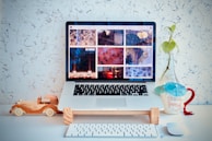 A laptop displaying a collection of images is placed on a desk against a textured white wall. In front of the laptop, there is a wooden toy car. To the right, a mug with an umbrella and a small plant in a glass bottle can be seen. A wireless keyboard and mouse are positioned on the desk.