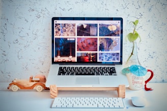A laptop displaying a collection of images is placed on a desk against a textured white wall. In front of the laptop, there is a wooden toy car. To the right, a mug with an umbrella and a small plant in a glass bottle can be seen. A wireless keyboard and mouse are positioned on the desk.