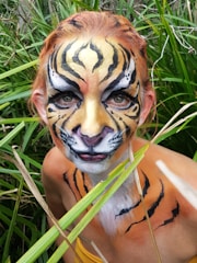 A person with their face and upper body painted like a tiger, surrounded by dense green foliage. The face is intricately detailed with yellow, black, and white paint that mimics a tiger's stripes. The background of tall grass adds to the jungle theme.