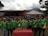 A group of young men wearing green sports jerseys with yellow accents are standing in a line, each displaying medals around their necks. In front of them is a table with several shiny trophies. Behind them, there is a small stage, a tent, and a mountain in the background.
