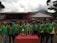 A group of young men wearing green sports jerseys with yellow accents are standing in a line, each displaying medals around their necks. In front of them is a table with several shiny trophies. Behind them, there is a small stage, a tent, and a mountain in the background.