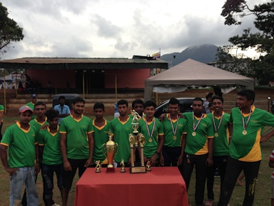 A group of young men wearing green sports jerseys with yellow accents are standing in a line, each displaying medals around their necks. In front of them is a table with several shiny trophies. Behind them, there is a small stage, a tent, and a mountain in the background.