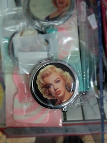 A round compact mirror features an image of a glamorous blonde woman with vintage-style makeup and earrings. The mirror is wrapped in plastic packaging and is surrounded by various other packaged items with similar designs.