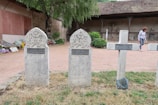 Three gravestones stand upright in a graveyard. Two of the gravestones are shaped with rounded tops and have Arabic inscriptions with a crescent moon and star symbol, while the third is a cross. There is a woman walking in the background near a stone building with a wooden roof. Flowers, trees, and bushes decorate the area around the gravestones.