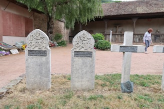 Three gravestones stand upright in a graveyard. Two of the gravestones are shaped with rounded tops and have Arabic inscriptions with a crescent moon and star symbol, while the third is a cross. There is a woman walking in the background near a stone building with a wooden roof. Flowers, trees, and bushes decorate the area around the gravestones.