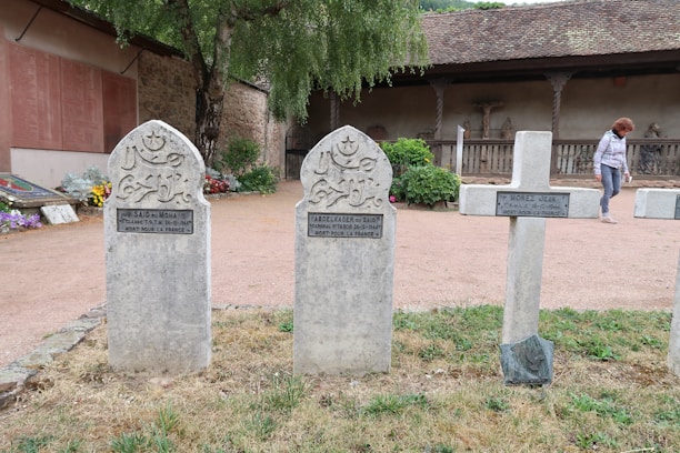 Three gravestones stand upright in a graveyard. Two of the gravestones are shaped with rounded tops and have Arabic inscriptions with a crescent moon and star symbol, while the third is a cross. There is a woman walking in the background near a stone building with a wooden roof. Flowers, trees, and bushes decorate the area around the gravestones.