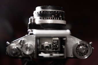 A close-up view of an analog camera, focusing on the lens and the top controls. Through the viewfinder, there's a reflection of another camera. The camera is dark with metallic components and intricate dials, giving it a classic, vintage appearance.