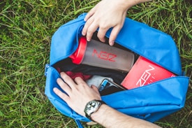 A person arranges items in a blue gym bag laid on green grass. Inside the bag, there are a protein shaker with 'ZEN' printed on it, a red box, likely a product package, and a smartphone with a blue casing. The person is wearing a watch on their left wrist.