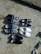 A variety of shoes arranged neatly on a rough, concrete surface. There are five pairs of casual footwear including slip-ons and sneakers. The shoes come in different designs and colors, such as blue with a horse logo, black, and white with a red and blue stripe. A person's foot in a sandal can be seen on the left edge of the image.