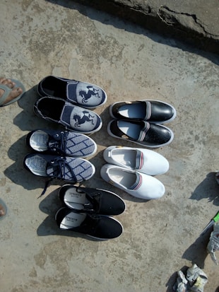 A variety of shoes arranged neatly on a rough, concrete surface. There are five pairs of casual footwear including slip-ons and sneakers. The shoes come in different designs and colors, such as blue with a horse logo, black, and white with a red and blue stripe. A person's foot in a sandal can be seen on the left edge of the image.