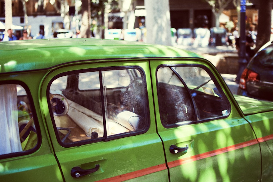 A vibrant green vintage car is parked on a sunlit street. Sunlight filters through the leaves, casting dappled shadows on the car's surface. The interior is partially visible through the window, revealing a rolled-up white item on the back seat. In the background, people and other parked cars can be seen, creating a lively urban scene.