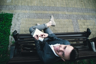 A person is sitting on a park bench writing in a notebook. They are dressed in a business-casual outfit, wearing a blazer and loafers. The bench is situated on a paved area surrounded by green foliage.